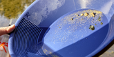 A close-up of a blue plastic gold pan being tilted over water, with gravel and sand inside the pan. A hand is visible holding the pan on the left side.
