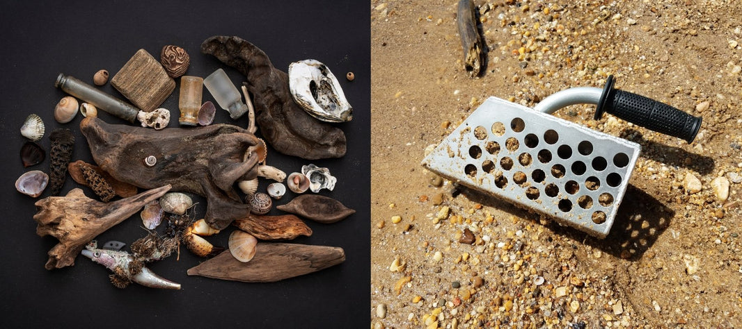 Beachcombing finds like driftwood, seashells, sea glass, and oyster shells arranged on a black background beside a metal sand scoop with a rubber grip resting on a wet, pebbly beach.