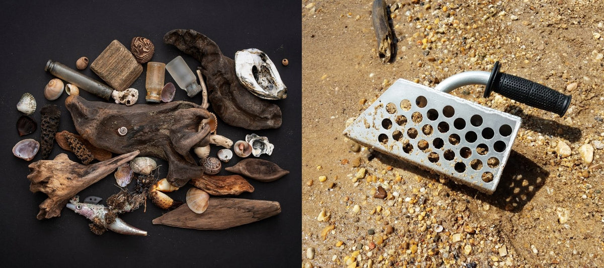 Beachcombing finds like driftwood, seashells, sea glass, and oyster shells arranged on a black background beside a metal sand scoop with a rubber grip resting on a wet, pebbly beach.
