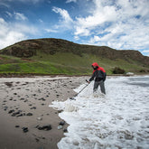 man on beach metal detecting in shallow water with a mountain in the background