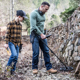 Two People metal detecting with the Nokta Score metal detector along a stone wall with trees in the background