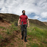 Man holding a manticore metal detector walking through a mountain Range with a cloudy sky 