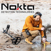 Man Using a pinpointer on a beach with a metal detector behind him With the Nokta Detection Technologies Logo