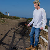 A man wearing a white sweatshirt and blue jeans stands outdoors by a wooden fence on a dirt path, holding a Quest  Q30 metal detector. He has a black wireless headphones on his ear.