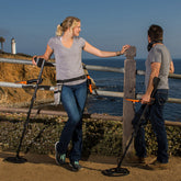 Two people standing outdoors by a coastal railing, each holding the Quest Q40 a black metal detector with orange accents. A lighthouse and ocean are visible in the background.