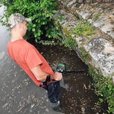 A man in an orange t-shirt and camouflage cap stands in shallow water holding a Teknetics EuroTek metal detector over a stone and grassy bank.
