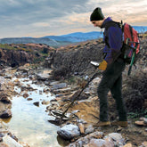 A person outdoors by a rocky stream using a Teknetics Gamma 6000 metal detector, wearing a dark beanie, purple jacket, dark pants, gloves, and a red backpack in a mountainous landscape.