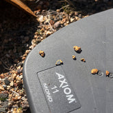 Close-up of a black Axiom 11 mono  coil on gravel, with a few small orange-brown objects scattered on the surface and the Axiom brand name visible.