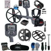 A metal detector bundle on a white background, including a gray and black metal detector with multiple control units, headphones, a battery charger, coil covers, gloves, a carrying bag, and small accessories. Visible branding includes MineLab and Serious Detecting.