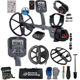 A metal detector kit laid out on a white background, including a gray and black metal detector with two coils, two handheld controllers, a pair of headphones, a battery charger, spare batteries, a carrying bag, gloves, a digging knife, a shovel, and other accessories. The detector and accessories show the NME / Serious Detecting branding.
