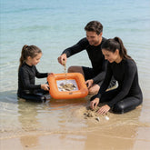 A family of three in black long-sleeve swimwear playing with a small orange inflatable sand tray on a sandy beach in shallow water.