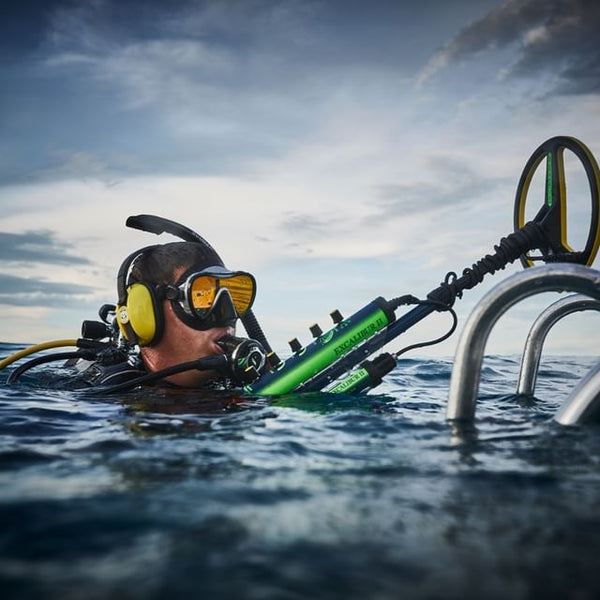 Scuba diver at the water’s surface using a Minelab Excalibur underwater metal detector near a metal ladder in open water.