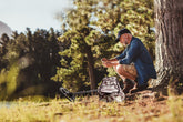 Man sitting by a tree with a metal detector in a forest setting