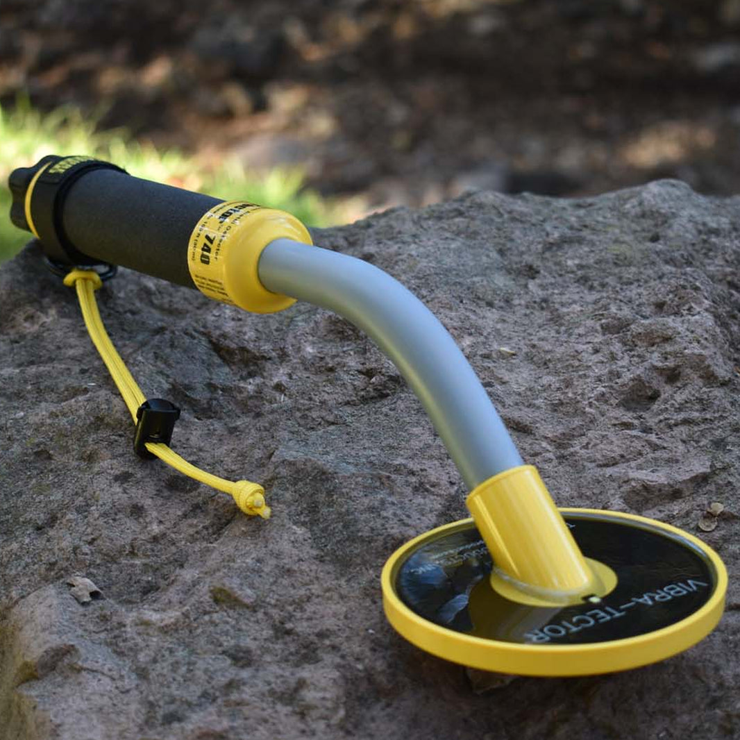 A handheld metal detector with a curved gray shaft and a yellow-and-black circular coil, resting on a large rock outdoors.