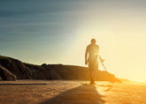 Person standing on a beach with a Nokta The Legend 2 Metal Detector and sunset in the background