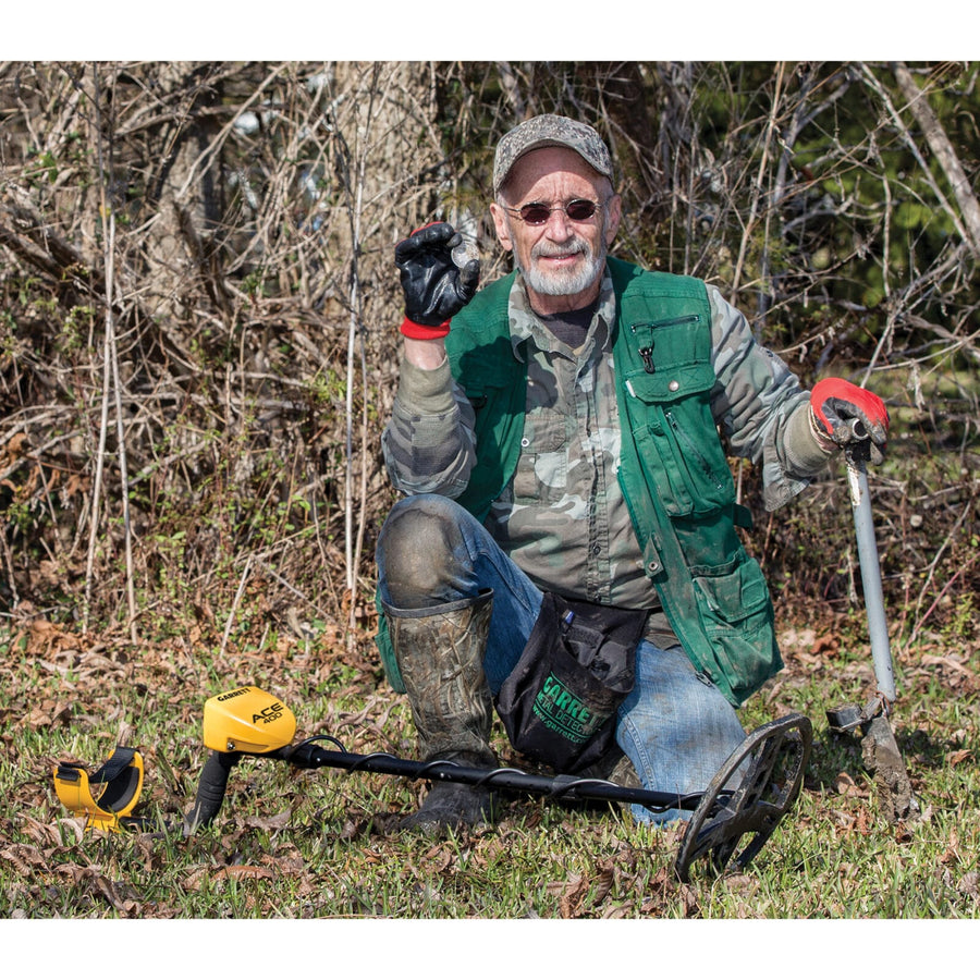 Detectorist kneeling in the woods beside a Garrett ACE 400 with a digger and recovered target