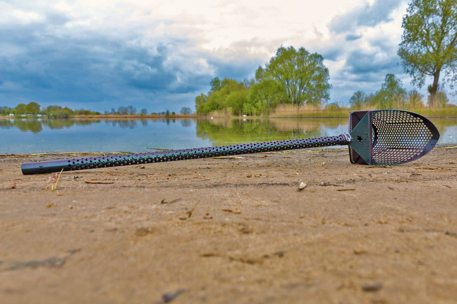 Motley Beach Sand Scoop with handle lying on a sandy lakeshore